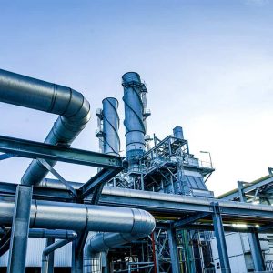 View of an industrial facility with large metal pipes, tall exhaust stacks, and structural frameworks against a clear blue sky—showcasing the role of AI in driving digital transformation and optimizing asset performance.