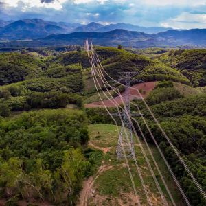 High-voltage transmission tower on a green hillside with several power lines stretching over a valley and distant mountains in view.