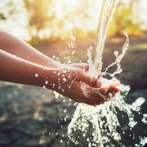 A person holds their hands under a stream of water, with water splashing and sunlight shining in the background—a reminder of the vital role compliance plays in ensuring clean, safe water through efficient water utilities.