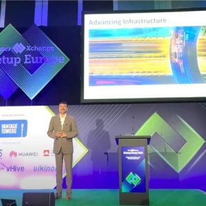 A man in a suit stands on stage giving a presentation titled "Advancing Infrastructure" at the TowerXchange Meetup Europe event. Sponsor logos are displayed on the stage background.