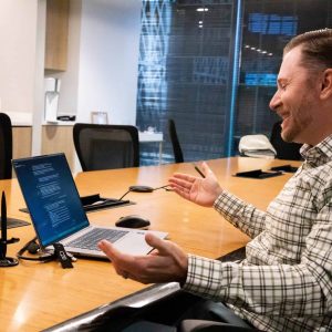A man sits at a conference table, gesturing and speaking during a video call on his laptop in a modern office setting.