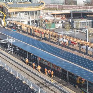 A group of construction workers wearing orange vests walk along train tracks beneath large arrays of solar panels at a modern transportation facility under construction, reflecting the engineering clarity championed by the Arcadis Rail Division.