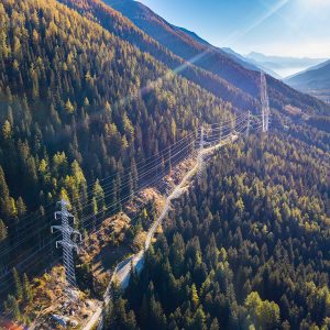 Aerial view of overhead power lines running through a forested mountain landscape under a clear sky, showcasing impressive power line engineering with distant valleys visible.