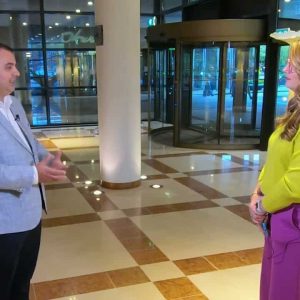 A man and a woman stand facing each other, discussing transportation value in a spacious, modern lobby with large windows and revolving doors.