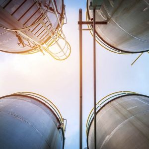 Four large industrial storage tanks viewed from below against a bright sky, with ladders and railings visible on the tanks' sides.