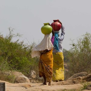 Two women walk on a rocky path carrying colorful water pots on their heads, while the natural background highlights how Digital Transformation can create solutions for remote communities.