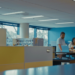 Four people stand and talk around a tall desk in a modern office with cubicles and large windows. A poster with a phone is visible on the left, highlighting the latest site design solutions like Bentley OpenSite+.