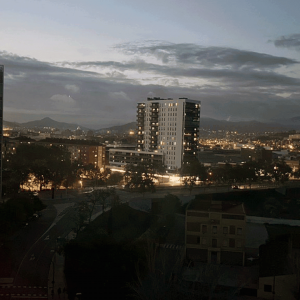 Cityscape at dusk with illuminated buildings, a central high-rise, dark tree silhouettes, and mountains in the background under a cloudy sky—the scene whispers stories of when the lights went out during the Iberian Blackout.