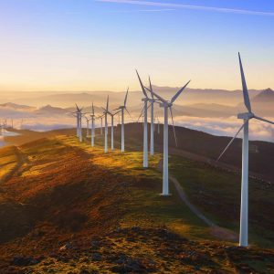 A row of wind turbines stands on a ridge at sunrise, with rolling hills and low clouds in the background.