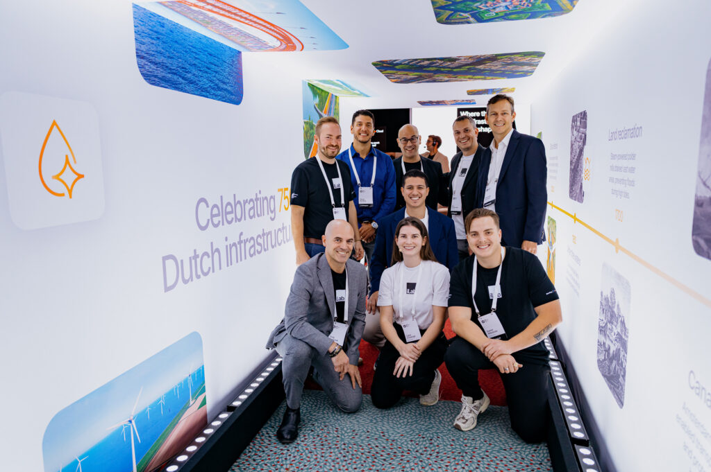 A group of nine people pose and smile inside a brightly lit exhibition tunnel with Dutch infrastructure displays on the walls.