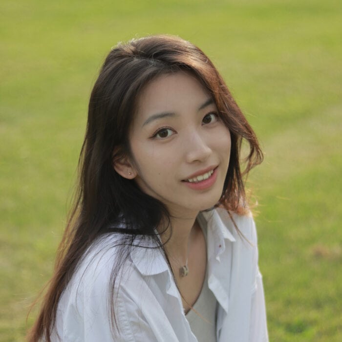 A woman with long hair and a white shirt smiles at the camera, sitting on a grassy field.