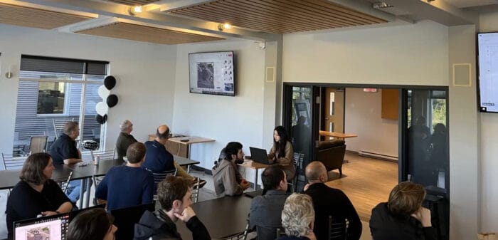 People sitting in a meeting room, watching a presentation displayed on screens.