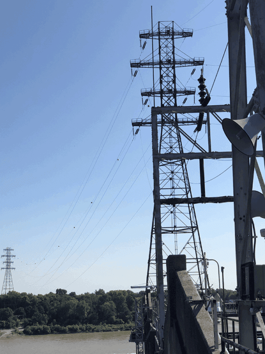Tall metal power lines tower beside a bridge over a river, with trees and a clear blue sky in the background.