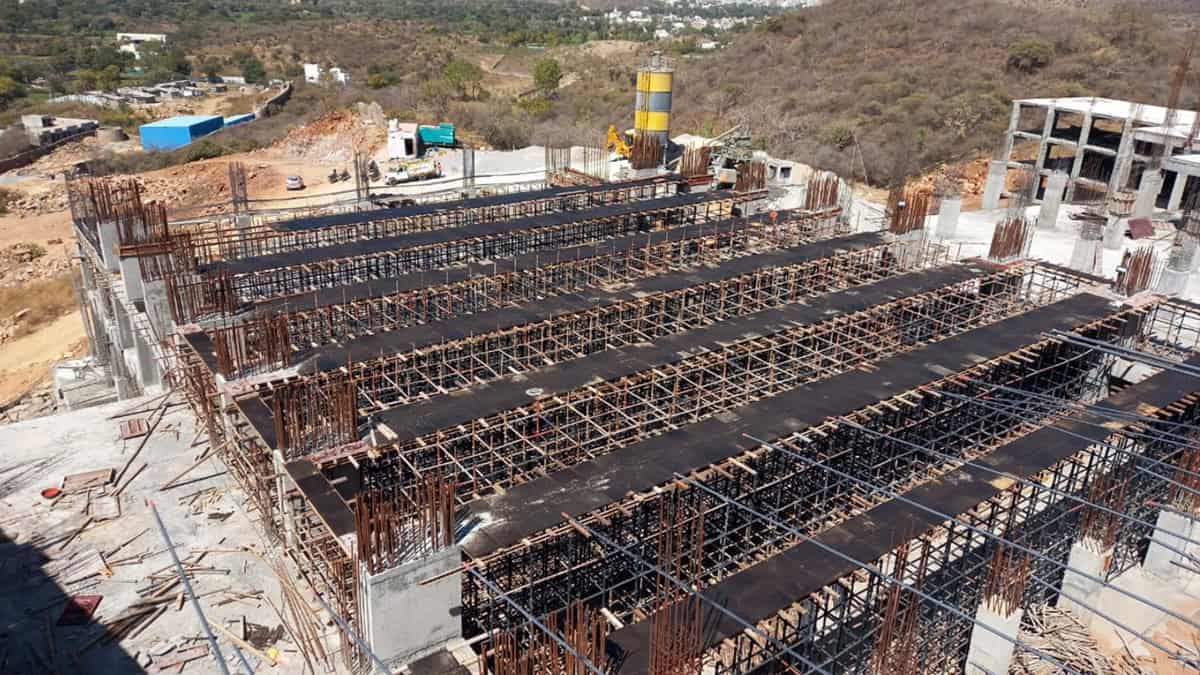 An under-construction building site by AVS ENGINEERS, showing multiple rows of steel scaffolding and formwork for concrete slabs, with hills and structures—including the Fairmont Palace Udaipur—visible in the background.