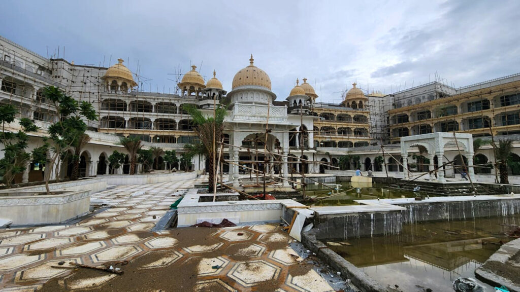 A large, ornate building with domes under construction by ISID STRUCTURES, surrounded by scaffolding, with an empty and unfinished fountain area in the foreground.