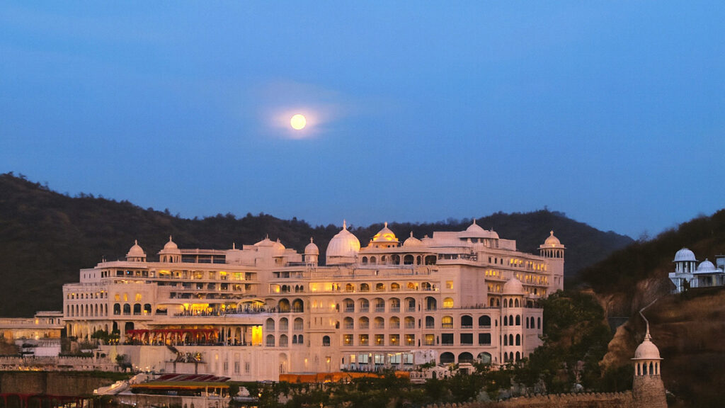 Large illuminated white building with domes and arches set against hills at dusk, with a full moon visible in the evening sky—this is the stunning Fairmont Palace Udaipur, a YII Winner designed with AVS ENGINEERS’ expertise.