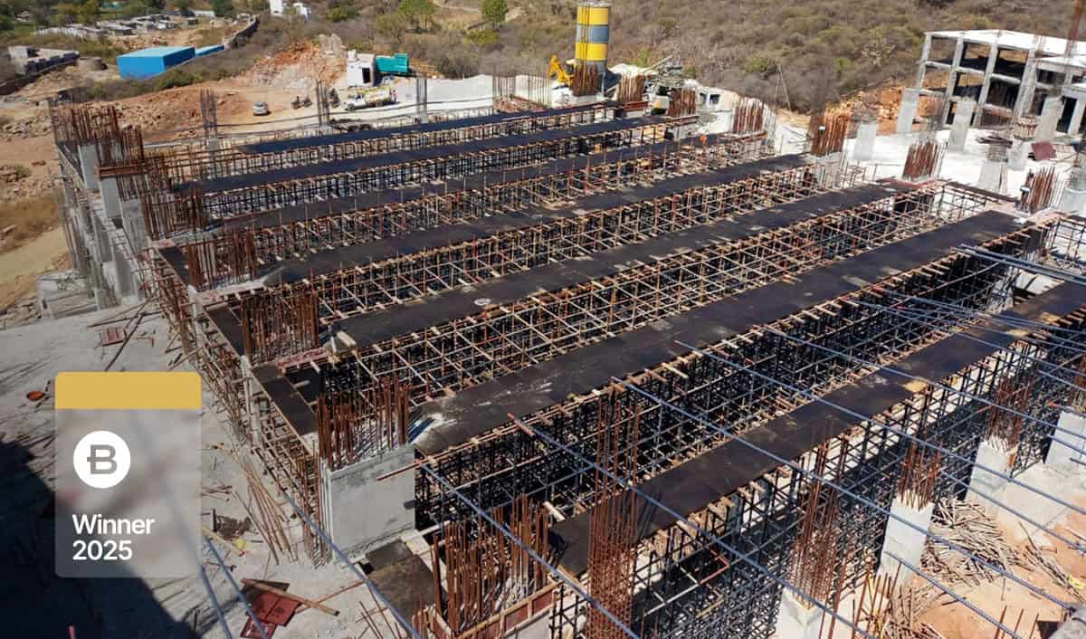 A construction site with concrete columns and steel scaffolding framework, showing early stages of building development at Fairmont Palace Udaipur, with hills and equipment in the background. A "Winner 2025" badge is overlaid.