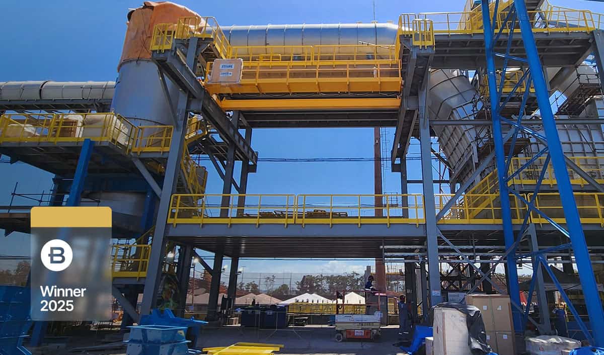 A large industrial facility, possibly a green briquette plant, with metal structures, pipes, and yellow railings under a clear sky; a “B Winner 2025” badge is visible in the lower left corner.