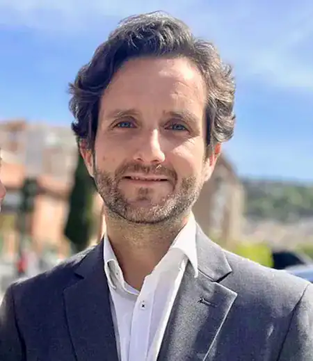 A man in a gray suit and white shirt stands outdoors on a sunny day, with buildings and trees blurred in the background.
