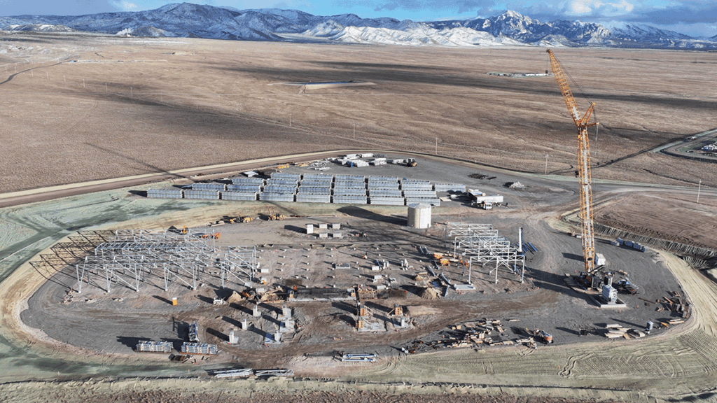 Aerial view of a construction site with steel framework, a tall crane, building materials, and mountains in the background.