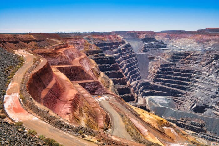 Large open-pit mine with terraced sides and winding roads, showing exposed layers of earth and rock under a clear blue sky.
