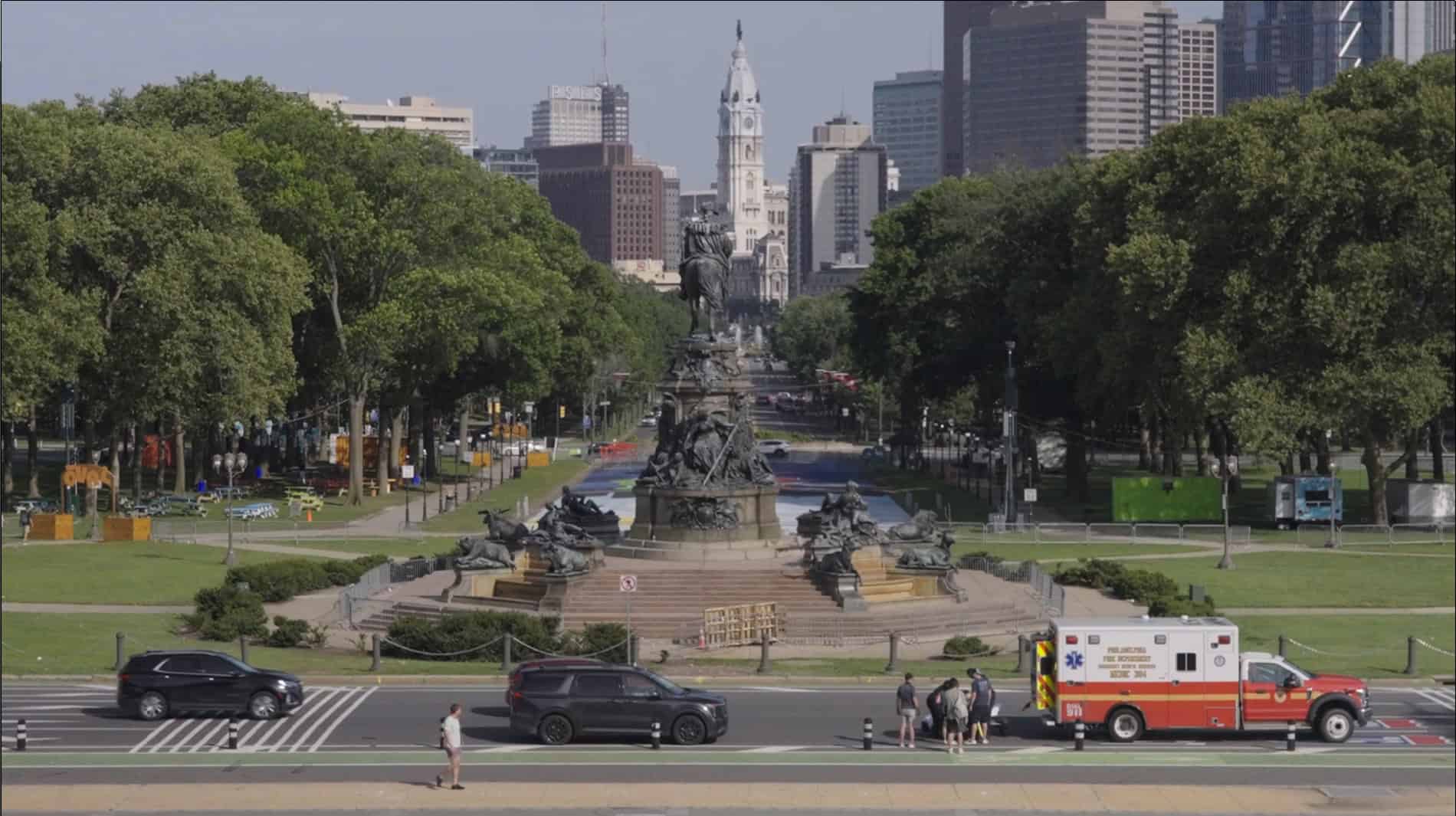 A view of the Washington Monument fountain in Philadelphia with City Hall in the background, trees lining the boulevard, vehicles, and an ambulance in the foreground.