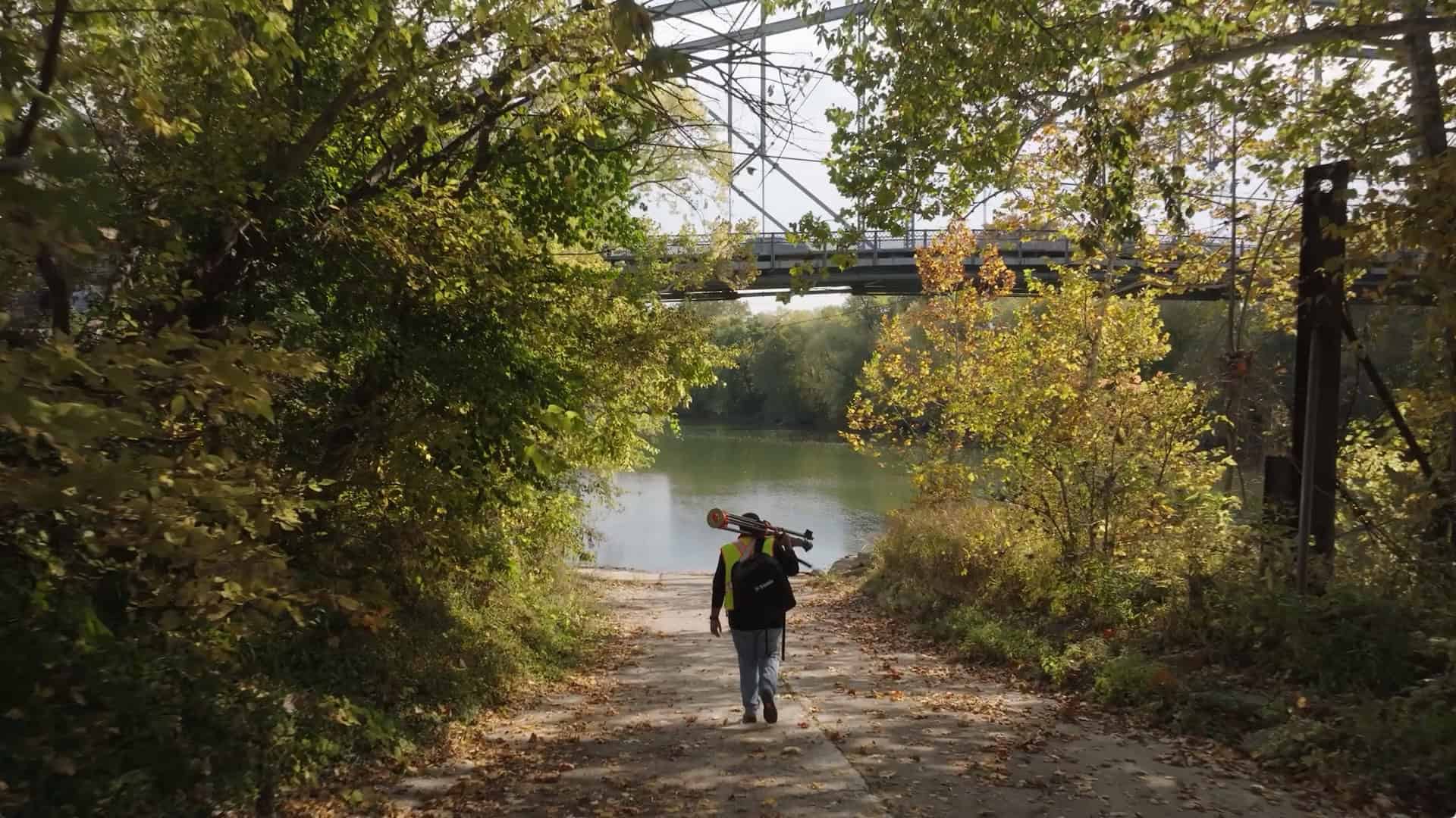 Person walking down a path toward a river, carrying a fishing rod. A bridge is visible in the background surrounded by trees with autumn foliage.