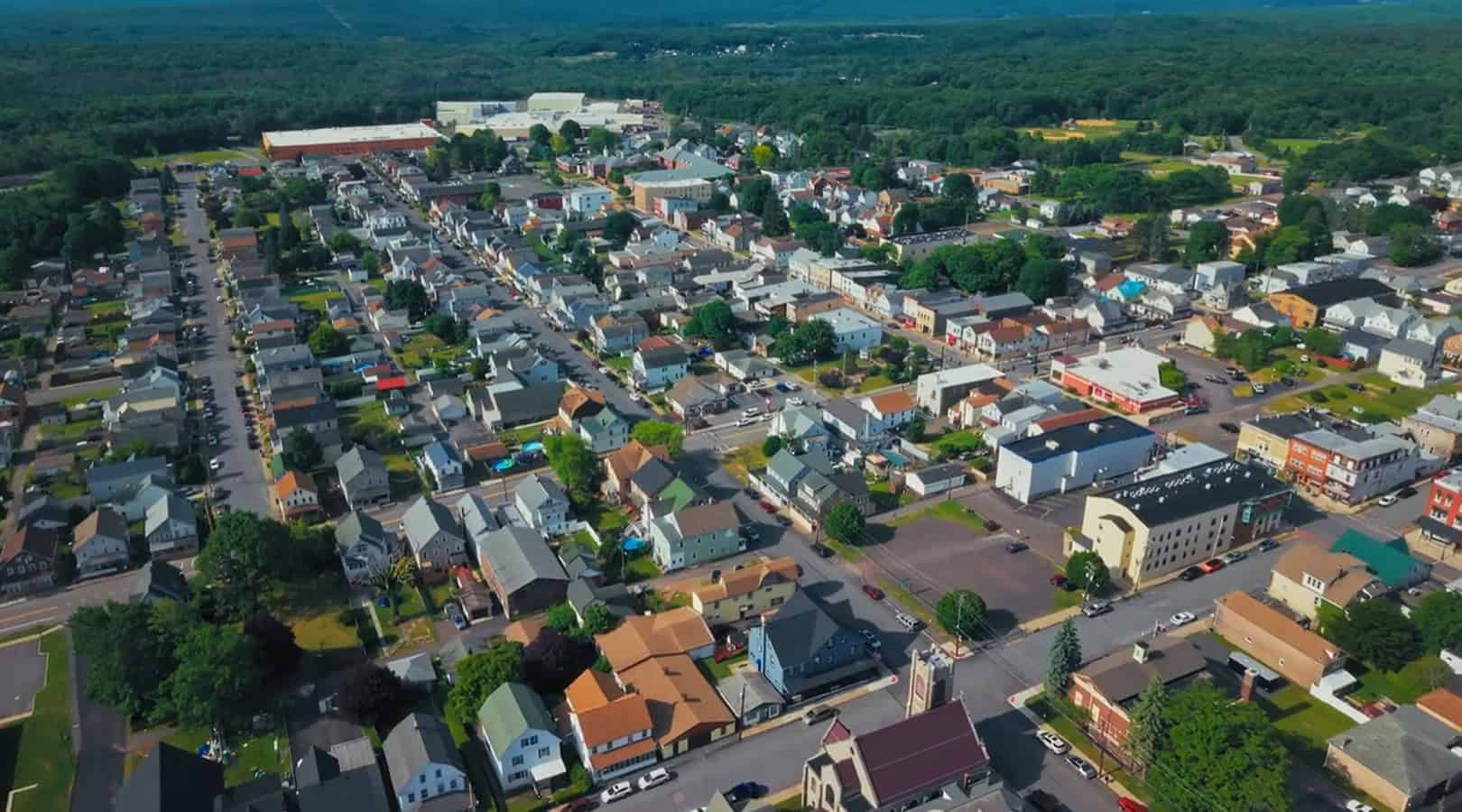 Aerial view of a small town with rows of houses, tree-lined streets, and various buildings surrounded by green, forested areas—showcasing how the Freeland Municipal Authority utilizes digital twins to monitor and manage water loss throughout the community.