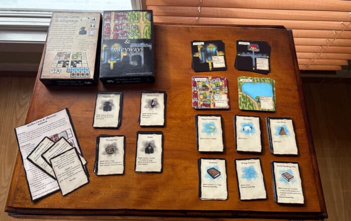 Board game components on a table, including cards, maps, instruction sheets, and box with "Glades, Caves & Alleyways" title.