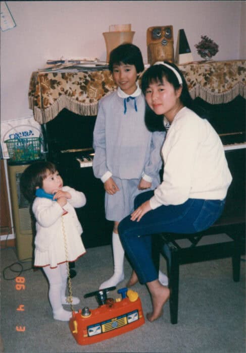 Three people are in a room; a toddler holding a toy phone, a young girl standing in a light blue dress, and another girl seated on a piano bench. A piano with a cover and decor items is in the background.