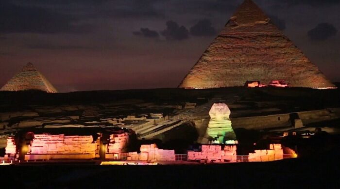 The Pyramids of Giza and the Sphinx illuminated by colorful lights at night, with a dark, cloudy sky in the background.