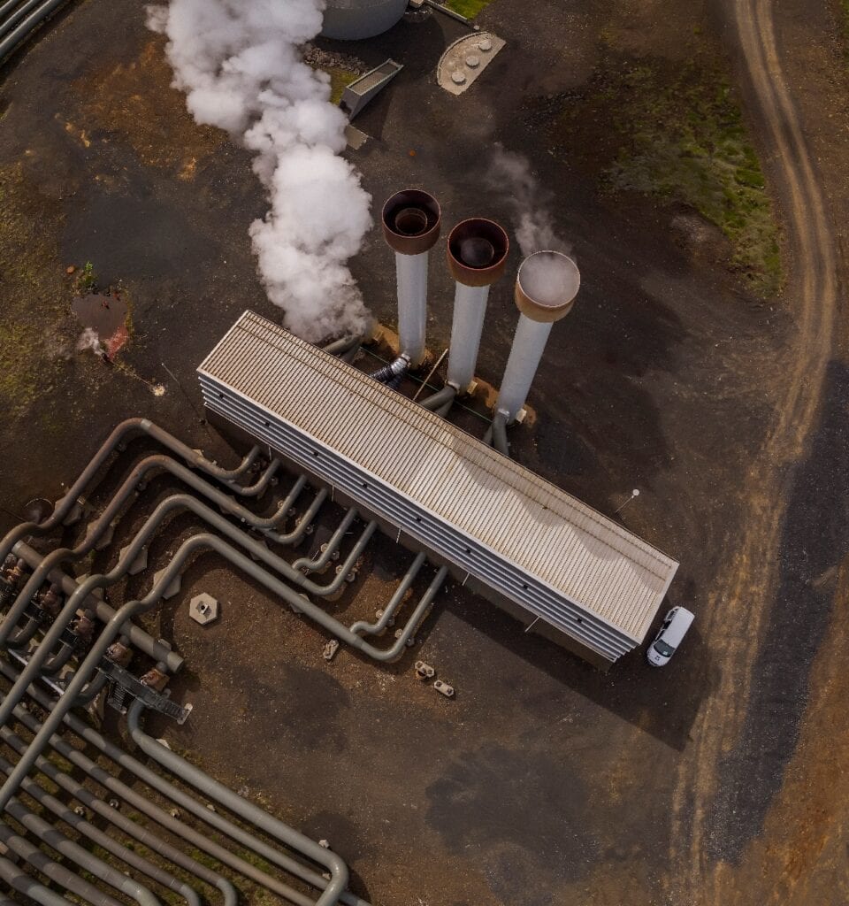 Aerial view of a geothermal energy facility with three smokestacks emitting white steam, surrounded by large pipes and a parked white vehicle.