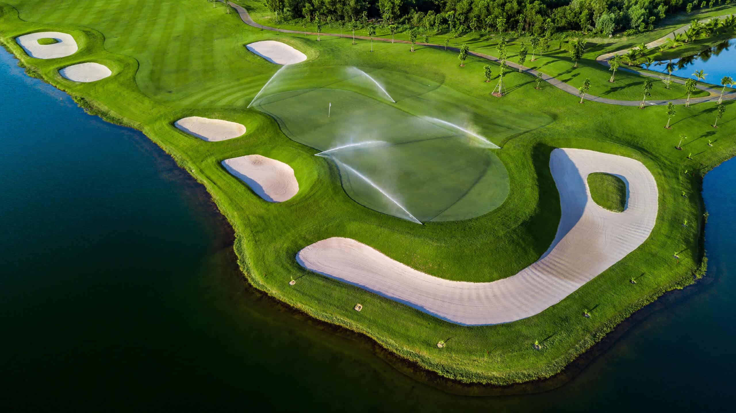 Aerial view of a golf course with water sprinklers on the green, surrounded by sand bunkers, a lake on the left, and trees in the background, seamlessly integrated into a nearby residential area.