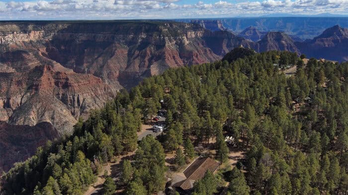 Aerial view of a forested area on the North Rim of the Grand Canyon with a few buildings and parked cars, surrounded by the expansive canyon landscape under a partly cloudy sky.