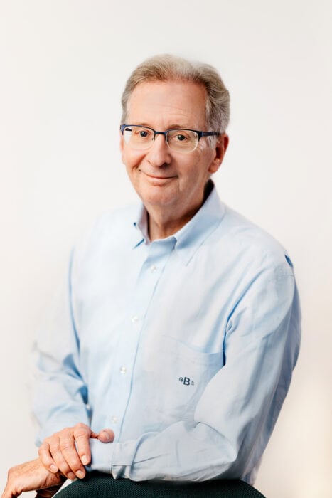 An older man with gray hair and glasses, wearing a light blue button-down shirt, sits facing the camera against a plain light background.
