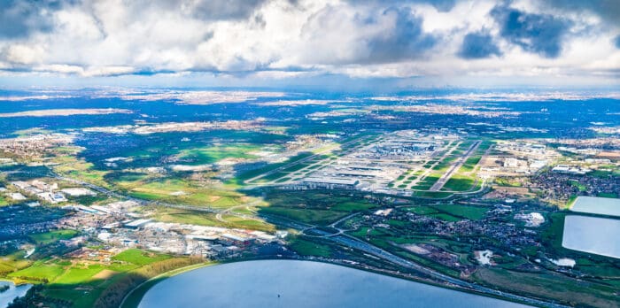 Aerial view of a large airport with multiple runways and terminals, surrounded by green fields, water, and scattered buildings under a partly cloudy sky.