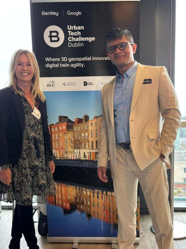 Two people stand in front of a banner for the Urban Tech Challenge Dublin, featuring Geospatial Innovation and event partners’ logos like WGIC and Bentley, with a cityscape photo in the background.