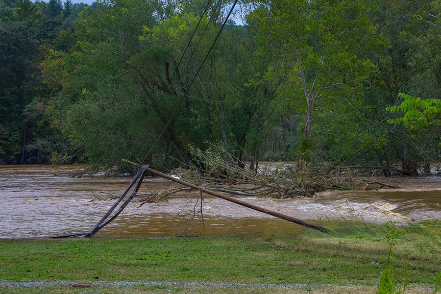 A broken utility pole leans over a swollen river with muddy water and debris after flooding, surrounded by trees and green grass.