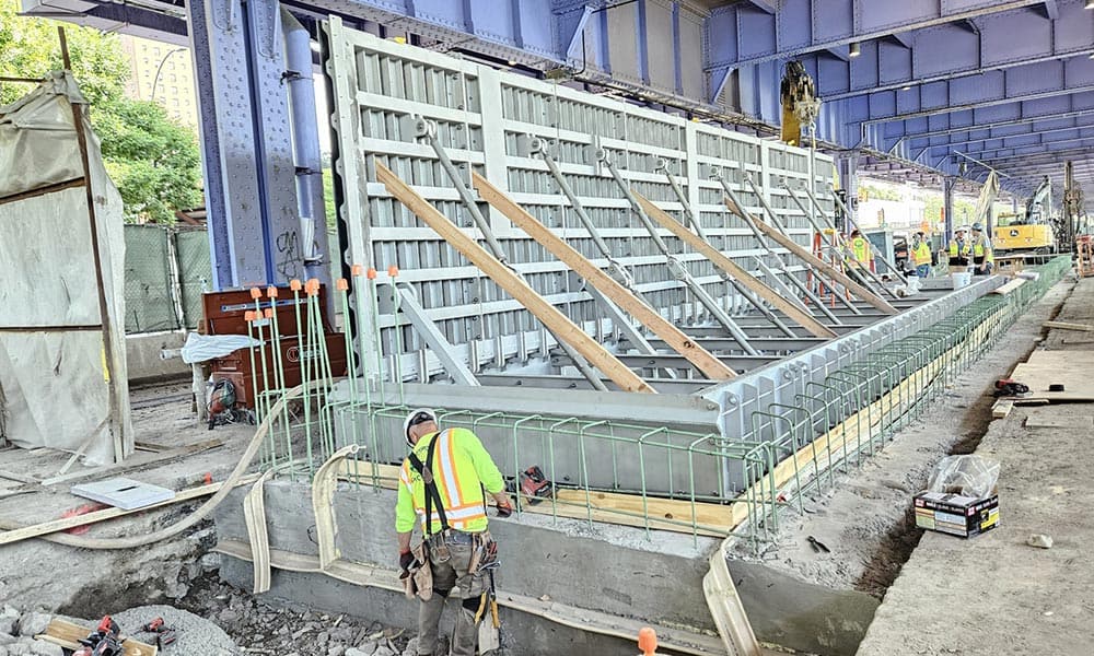 Construction workers in NYC reinforce a concrete structure under a large metal bridge, using wooden supports and metal rebar. Tools and equipment around the site highlight efforts toward climate resilience.