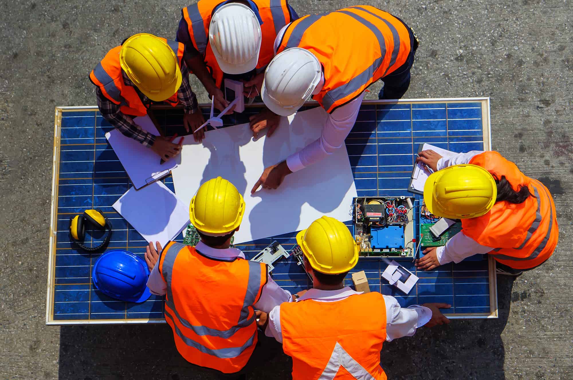 Construction workers in high visibility vests and safety helmets discussing plans over a blueprint with a solar panel nearby.