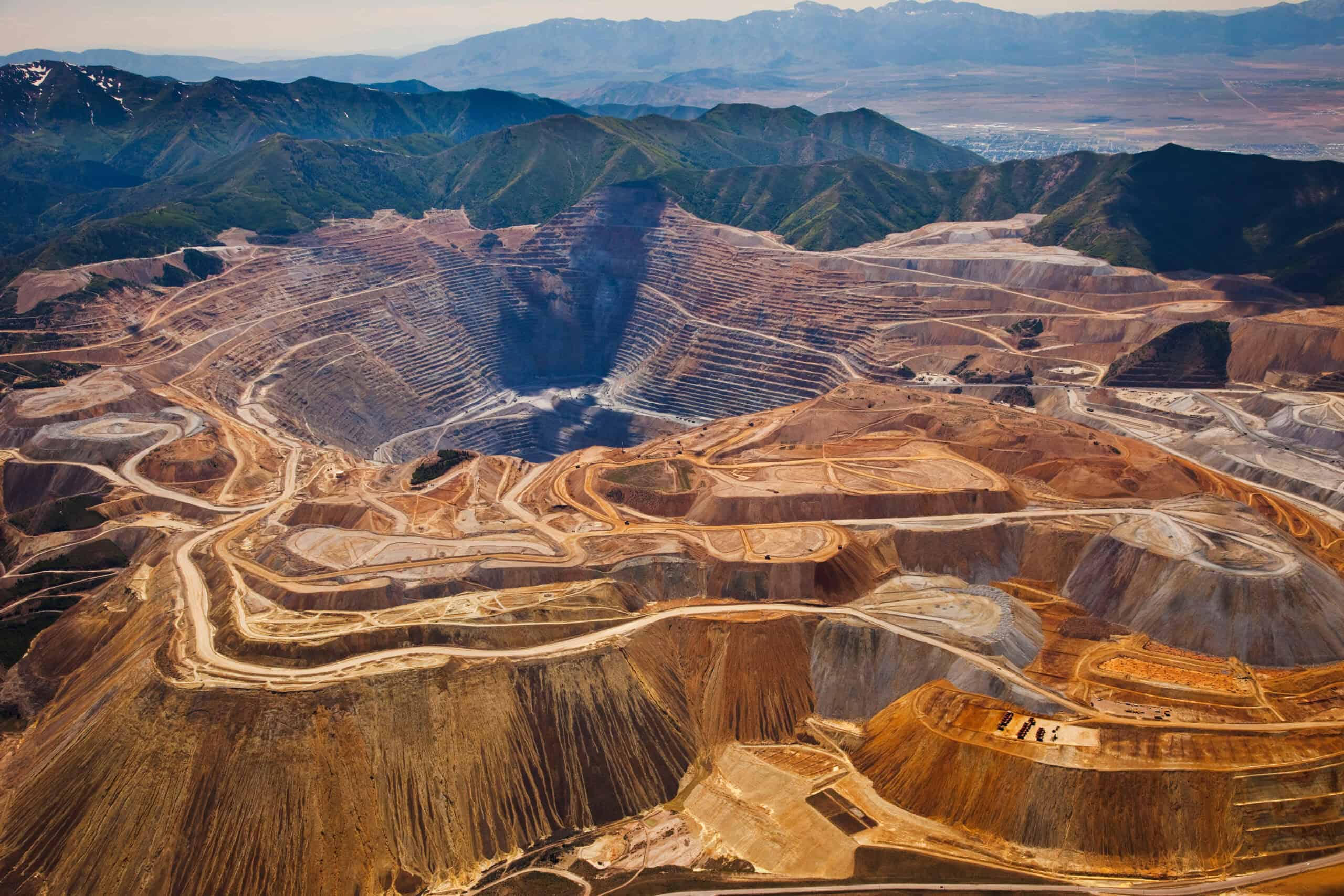 Aerial view of a large open-pit mine with terraced slopes, winding roads, and surrounding mountainous landscape.