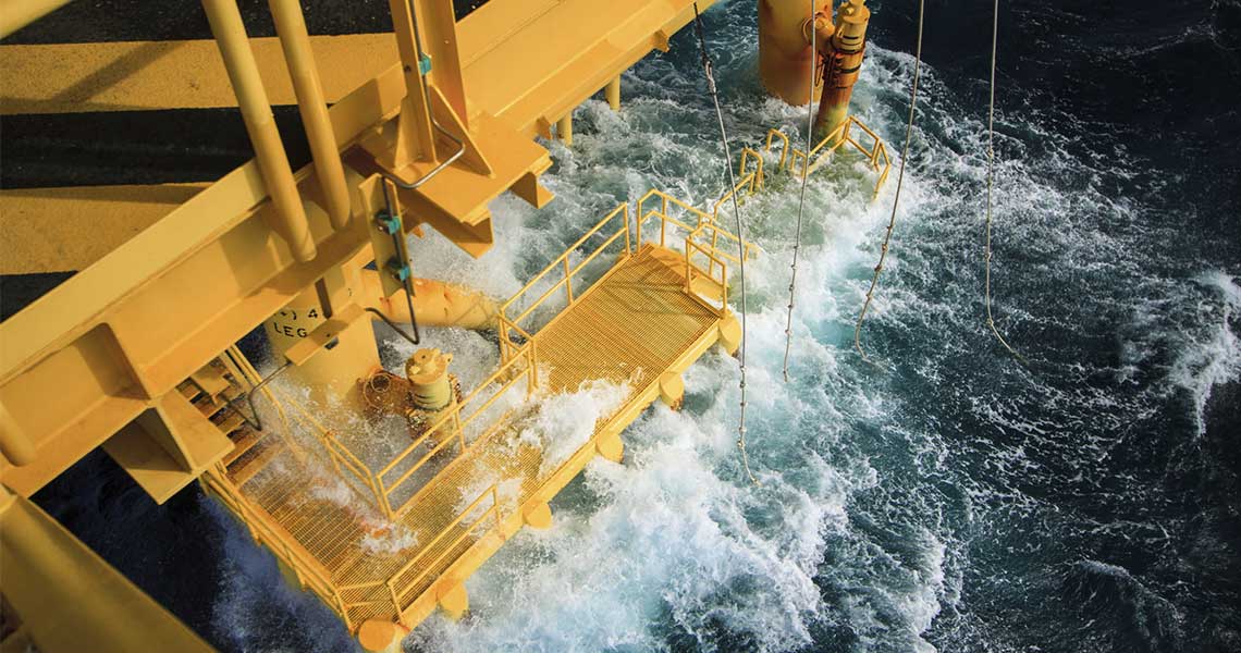 A yellow offshore oil platform structure extends over rough ocean waves, with water splashing onto the metal deck and equipment.