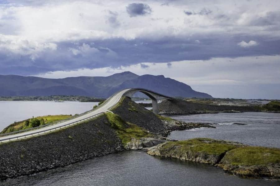Curving bridge over coastal waters against a backdrop of mountains and cloudy skies.