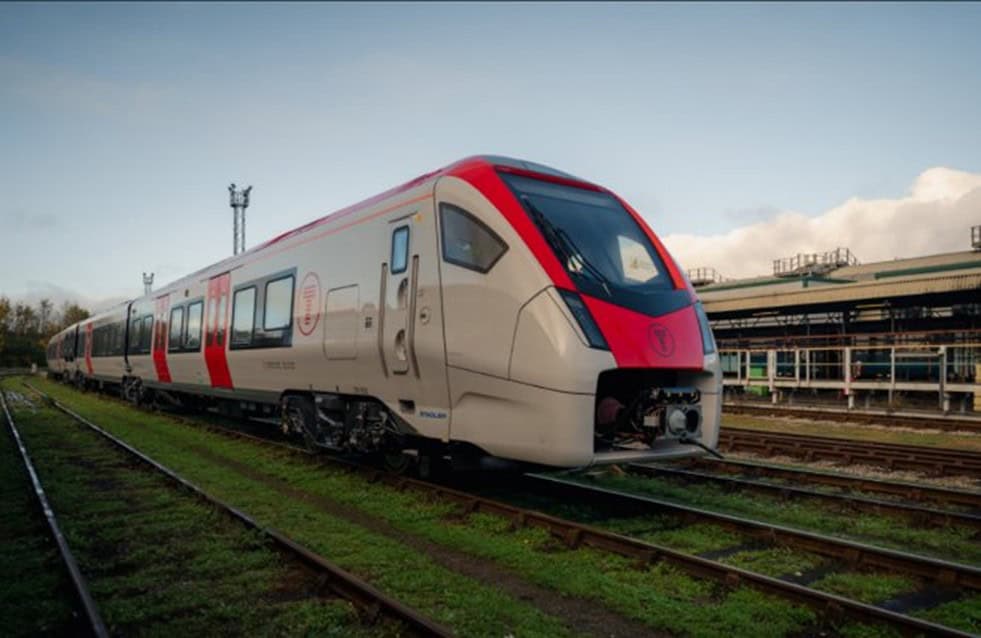 A modern passenger train with red and white livery is stationary on railway tracks beside an industrial building under a partly cloudy sky.