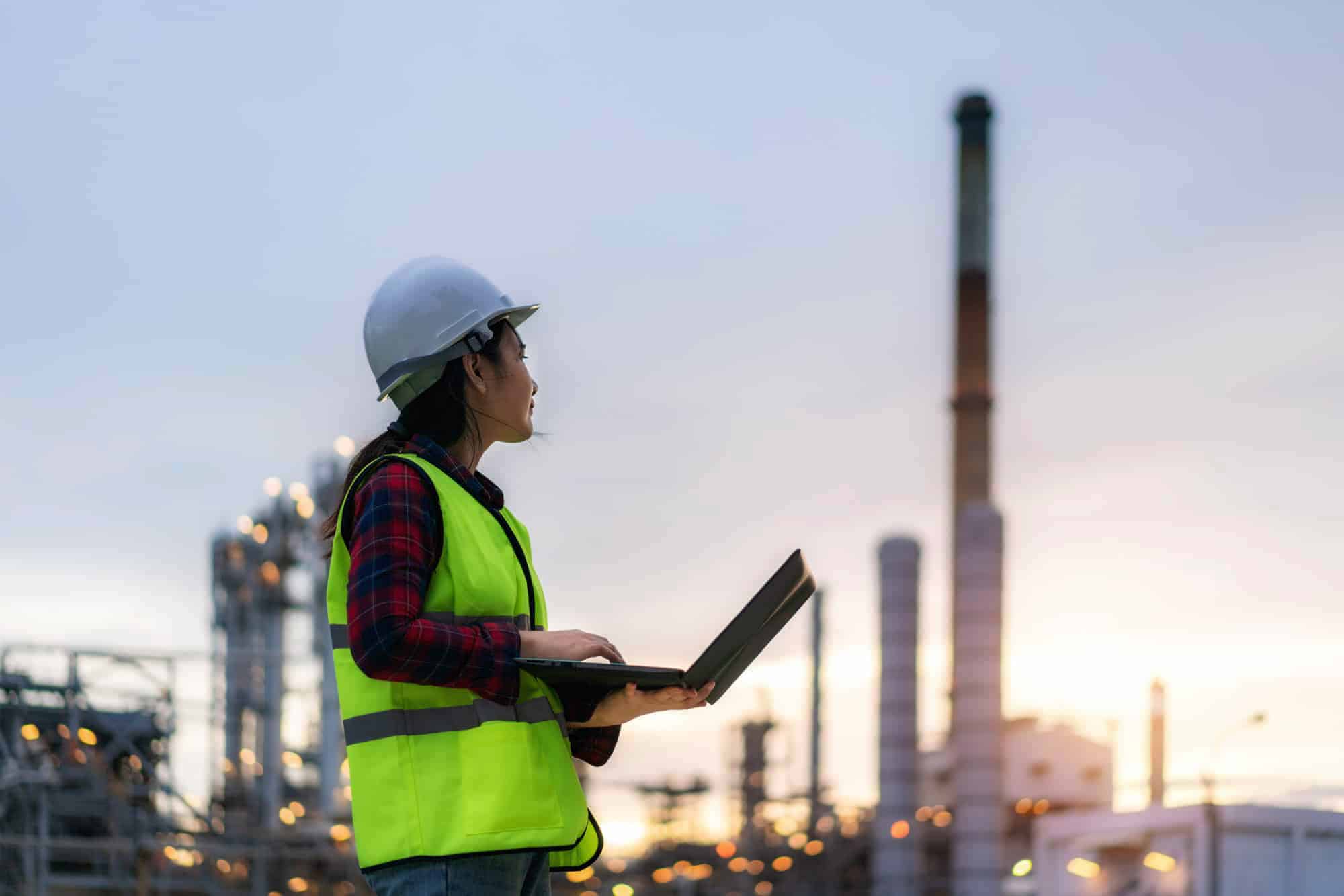 Female engineer with laptop at industrial site during dusk.