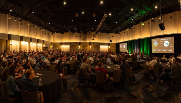 The 2025 Bentley Civil User Conference features a large conference room filled with attendees at round tables, all focused on the stage where multiple screens showcase the latest engineering insights and presentations.