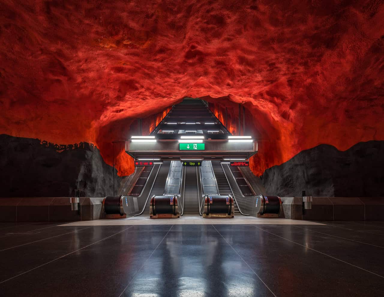 A modern underground station with a striking red, rock-like cave ceiling, illuminated escalators, and green emergency exit signs showcases the elegance of advanced underground construction techniques.