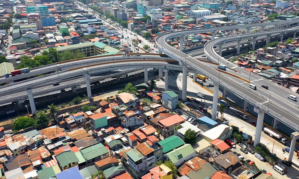 Aerial view of an elevated highway interchange, a testament to engineering firms’ expertise, passing above densely packed residential buildings in a thriving urban area.