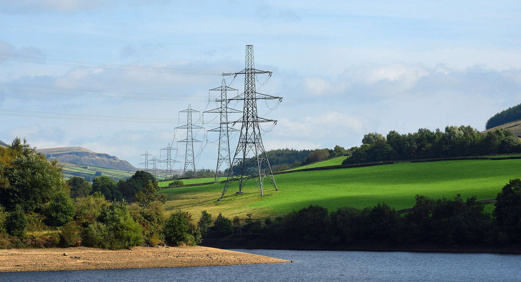 Electricity pylons and power lines cross a green landscape with trees, hills, and a body of water under a partly cloudy sky.