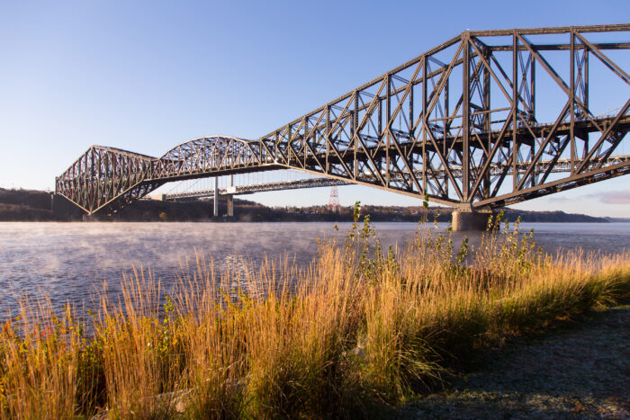 A large steel cantilever bridge spans over a wide river, with tall grass in the foreground and a clear blue sky overhead.